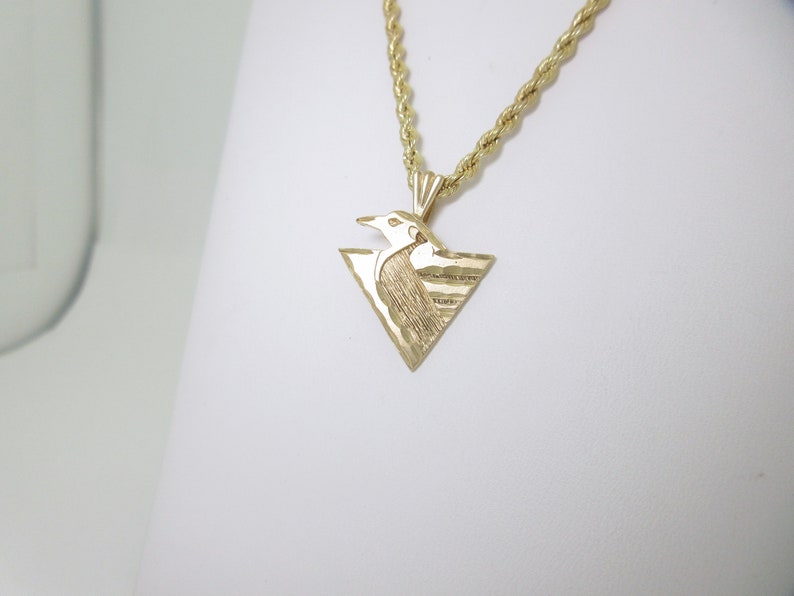 Puede incluir: Un collar de cadena de oro con un colgante triangular que presenta un dise&ntilde;o de p&aacute;jaro estilizado.