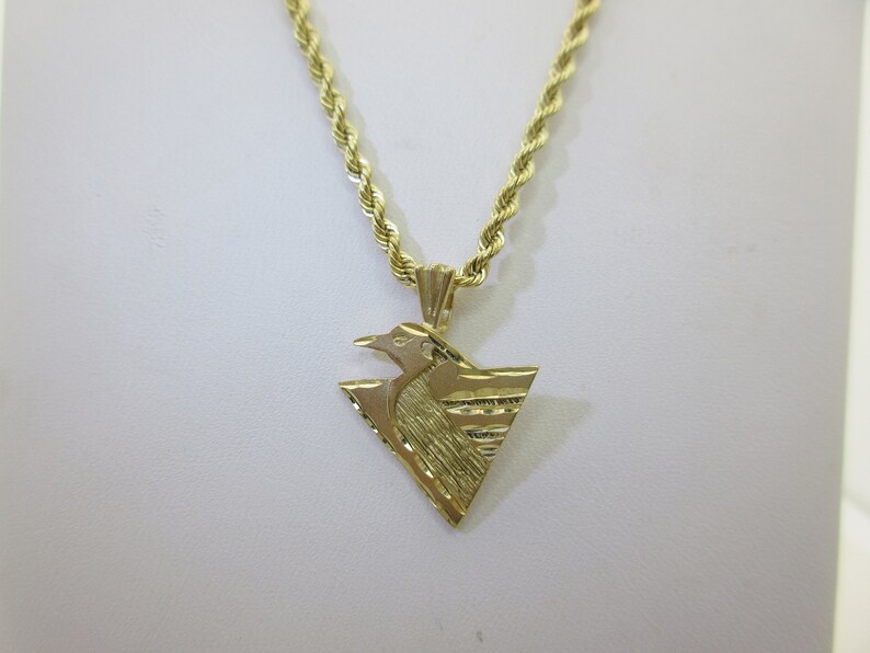 Puede incluir: Un collar de cadena de oro con un colgante que presenta un dise&ntilde;o de p&aacute;jaro estilizado. El colgante tiene forma de tri&aacute;ngulo con una superficie texturizada.