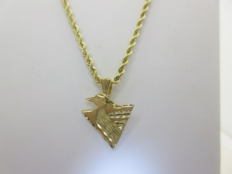 Puede incluir: Un collar de cadena de oro con un colgante que presenta un dise&ntilde;o de p&aacute;jaro estilizado. El colgante tiene forma triangular y una superficie texturizada.
