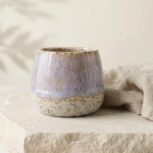 Puede incluir: Una taza de cerámica con un esmalte liso y brillante en tonos lavanda y azul claro en la parte superior y un acabado texturizado y moteado en color blanco roto en la base. El recipiente tiene una forma redondeada sin asa.