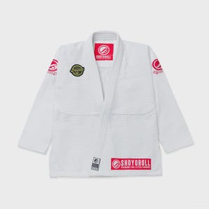 Peut inclure: Une veste de kimono d'arts martiaux blanche avec des logos Shoyoroll rouges sur le col, les épaules et le bas. Un écusson noir circulaire avec du texte doré est sur la poitrine droite. Le vêtement a des manches longues et un col renforcé.