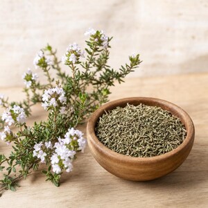 May include: A small wooden bowl filled with dried green thyme leaves next to a sprig of fresh thyme featuring tiny light purple flowers. The dried herb is finely chopped and textured, while the fresh plant has thin stems and small leaves.
