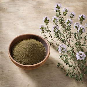 May include: A small, round, dark brown wooden bowl filled with a mound of finely ground, olive-green herbal powder, positioned next to a sprig of fresh thyme featuring thin green stems and clusters of small, light purple, bell-shaped flowers.