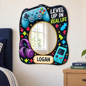 May include: A wall-mounted mirror with a black frame featuring colorful gaming graphics including a blue controller, purple headphones, a handheld console, and pixelated shapes. The text reads LEVEL UP IN REAL LIFE and the name LOGAN on a plaque.