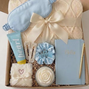 Op de afbeelding: Een geschenkdoos met een lichtblauw satijnen slaapmasker, een crèmekleurige kabelgebreide deken met een lint, een tube Pearl handcrème, twee witte haarclips, een lichtblauwe scrunchie, een pluizig wit vierkant doekje met een hartje, een roosvormige kaars, een lichtblauw notitieboek met de tekst Notes en een gouden pen.
