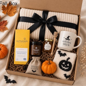 Könnte beinhalten: Eine Geschenkbox mit einer cremefarbenen, gerippten Strickdecke mit schwarzer Schleife, einer Moonstruck Milchschokolade mit gesalzenem Toffee, einer Sandalwood Pumpkin Kerze, einem Honigglas mit Holzlöffel, einer Witches Brew Tasse und flauschigen Socken.