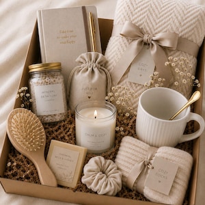 Puede incluir: Una caja de regalo con la etiqueta JOY BOX que contiene una manta beige de espiga, un cuaderno con un bolígrafo dorado, un frasco de sales de baño, una pequeña bolsa, una vela encendida, una taza blanca con cuchara dorada, un cepillo de madera, jabón y calcetines.