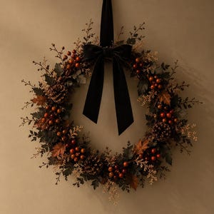 Può includere: Una ghirlanda circolare composta da fogliame verde scuro, pigne marroni e grappoli di piccole bacche rosse. La ghirlanda è decorata con un grande fiocco in velluto nero legato in alto, con due lunghi nastri che scendono dal centro.