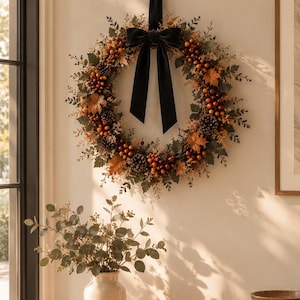 Peut inclure: Une couronne d'automne circulaire composée de feuilles d'érable artificielles orange et brunes, de pommes de pin et de petites baies, agrémentée d'un grand nœud en velours noir. La couronne est suspendue par un ruban de velours noir.