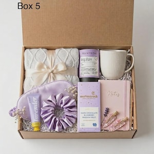 Op de afbeelding: Een geschenkdoos met het label Box 5 bevat een grijze kabelgebreide deken met een crèmekleurig lint, een witte geribbelde mok, een lavendelkleurig slaapmasker, een paarse scrunchie, een tube lavendelcrème, Moonstruck-chocolade en een roze notitieboek.