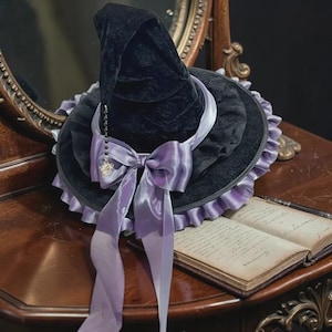 May include: A black velvet witch hat featuring a wide brim with a ruffled lavender trim. A large lavender satin bow is tied around the base, accented by a dangling silver-toned chain with a small star charm hanging from the pointed crown.