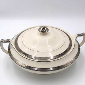 May include: A round, cream-colored ceramic serving dish with a matching lid and two side handles. The dish features a metallic silver band around the lid and the base, decorated with a subtle, repeating leaf-like pattern along the edges.