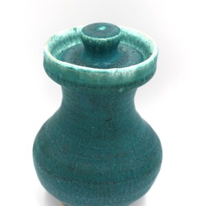 May include: A small, bulbous ceramic jar with a vibrant teal glaze and a matching lid. The jar features a narrow neck that flares out into a wide, circular rim. The glaze has a textured, mottled appearance with lighter white highlights.