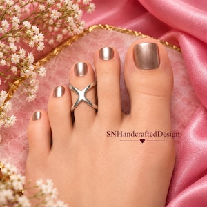 Puede incluir: Un anillo para el dedo del pie de metal plateado con un diseño simple en forma de X, colocado en el segundo dedo. Las uñas de los pies están pintadas con un esmalte color bronce metálico. El texto SNHandcraftedDesign aparece abajo a la derecha.