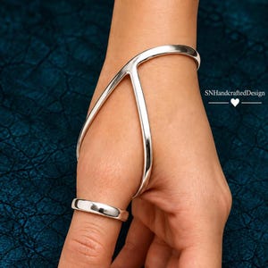 May include: A polished silver hand bracelet featuring a smooth, curved metal band that wraps around the wrist and connects to a simple ring encircling the thumb. The design includes the text SNHandcraftedDesign with a heart icon.