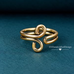May include: A gold-toned metal ring featuring an open, bypass-style design with two curved, hook-like ends that face opposite directions. The band has a smooth, polished finish. The text SNHandcraftedDesign is visible in the lower right corner.