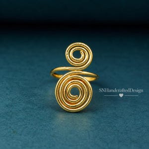 Può includere: Un anello color oro caratterizzato da due prominenti disegni a spirale collegati da una sottile fascia. La superficie metallica è liscia e riflettente. Il testo SNHandcraftedDesign con una piccola icona a forma di cuore è visibile a destra.