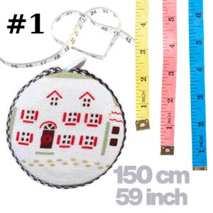 Op de afbeelding: Een rond oprolbaar meetlint met een witte stoffen behuizing voorzien van een kruissteekpatroon van huizen in rood en bruin. Het lint meet 150 cm of 59 inch. Drie extra meetlinten in geel, roze en blauw zijn ook zichtbaar.