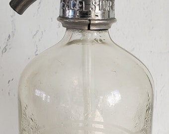 May include: A vintage glass soda siphon with a chrome top and handle. The siphon has the words "Excelsior Store" printed on the top. The bottle is clear and has a textured surface.