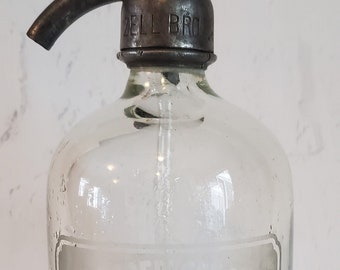 May include: A clear glass bottle with a metal pump top. The bottle has a label that reads "Alex. Bell & Sons, Chemists, Obban, Established 1846." The label also has a trade mark with a deer inside.