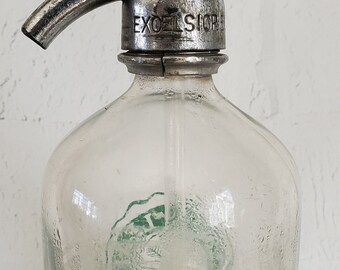 May include: A vintage glass bottle with a metal top and a silver lever. The bottle is clear and has a green design on the front. The metal top has the word "EXCELSIOR" embossed on it.