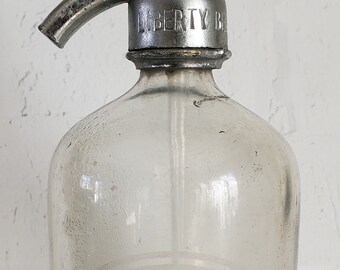 May include: A clear glass bottle with a silver metal top and a spout. The bottle is labelled "Frank Ewing Bottler" and "Contents 26 fl oz".