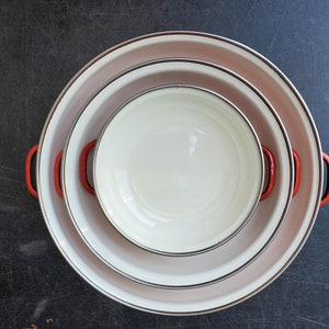 May include: Three nesting bowls with white enamel interiors and red and white rims. The bowls are stacked on a dark gray surface.