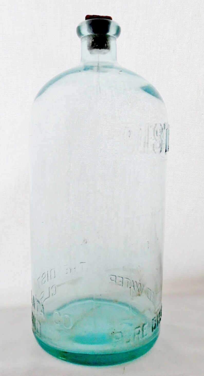 May include: A clear glass bottle with a cork stopper. The bottle is labeled "Pure Distilled Water" and "The Distilled Water Co."