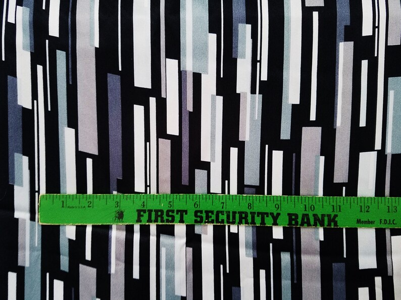 May include: Black fabric with a geometric pattern of white, gray, and light blue rectangles. A green ruler with the text "FIRST SECURITY BANK" is placed on top of the fabric.