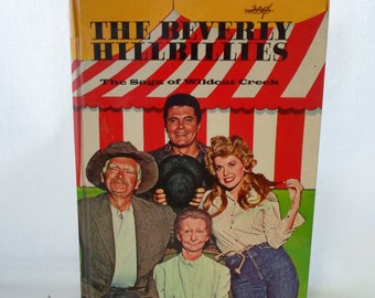Può includere: Una copertina di libro vintage con il cast della serie televisiva "The Beverly Hillbillies." La copertina presenta una tenda a strisce rosse e bianche con il titolo "The Beverly Hillbillies" in lettere gialle. Il sottotitolo "The Saga of Wildcat Creek" è stampato in lettere nere sotto il titolo. La copertina presenta quattro attori: un uomo con un cappello da cowboy, un uomo con una camicia nera, una donna con una camicia a quadri e una donna con una camicia bianca.