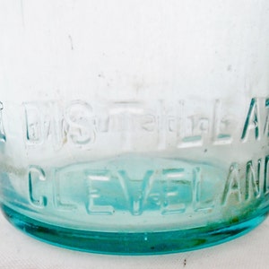 May include: Close-up of a clear glass bottle with a blue tint. The bottom of the bottle is embossed with the text "The Eagle A.T.A. Cleveland".