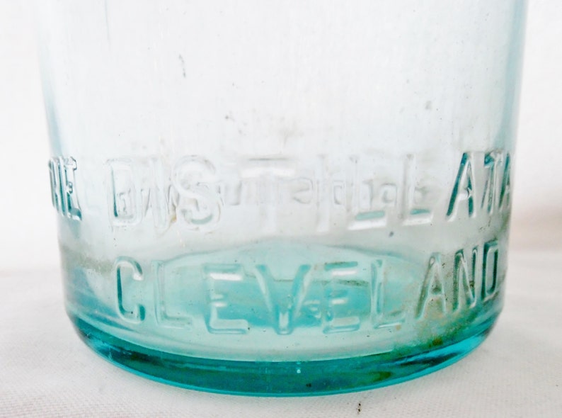 May include: Close-up of a clear glass bottle with a blue tint. The bottom of the bottle is embossed with the text "The Eagle A.T.A. Cleveland".