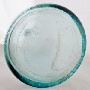 May include: The bottom of a clear glass bottle with a teal green rim. The bottle has a circular embossed mark on the bottom.