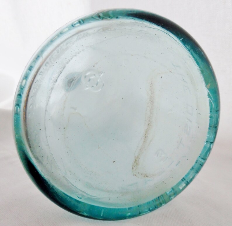 May include: The bottom of a clear glass bottle with a teal green rim. The bottle has a circular embossed mark on the bottom.