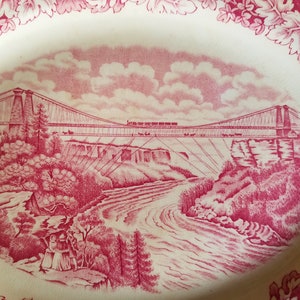 May include: A vintage oval ceramic plate with a pink floral border. The centre features a red and white illustration of a suspension bridge over a river with a train crossing it.