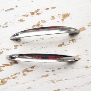 May include: Two silver-toned metal drawer pulls with a curved shape. The pulls are on a white, distressed wooden surface.