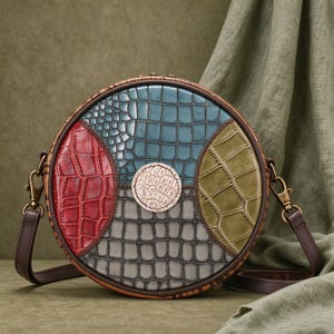 May include: A round crossbody bag featuring a multi-colored crocodile-embossed pattern in red, blue, olive green, and gray. The bag has a central circular patch, dark brown leather trim, and matching brown adjustable shoulder straps.