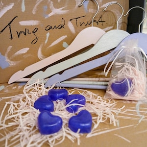Può includere: Tre grucce in legno nei colori rosa pastello, verde salvia e lavanda, ciascuna con un gancio in metallo argentato. Sotto si trovano cinque piccoli oggetti blu scuro a forma di cuore su strisce di carta rosa chiaro, con un cuore in un sacchetto.