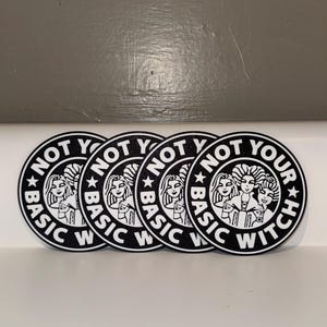 May include: Four circular black and white coasters featuring a graphic illustration of three women with elaborate hairstyles. The text "NOT YOUR BASIC WITCH" is printed in a circular border around the central image, with stars separating the words.