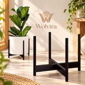 May include: A black wooden plant stand with a cross-shaped base and four vertical supports. In the background, a white cylindrical planter holds a green fiddle leaf fig plant. The logo Wolvira is visible in the center of the image.