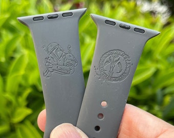 May include: Two gray silicone Apple Watch bands with engraved designs. One band features a Star Wars character, and the other band features a logo.