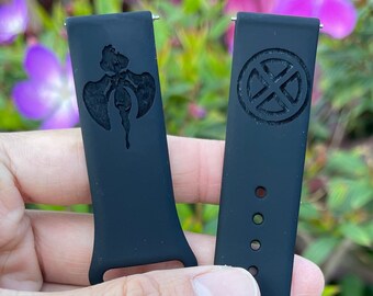 Op de afbeelding: Twee zwarte horlogebandjes met verschillende ontwerpen. Eén bandje heeft een afbeelding van een gestileerd figuur met vleugels. Het andere bandje heeft een afbeelding van een cirkel met een X erin.