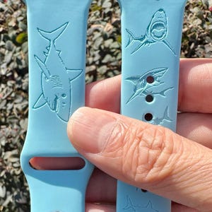May include: Light blue watch bands with shark designs. The bands feature multiple shark illustrations in a repeating pattern. The bands are made of a flexible material and are designed to fit a smartwatch.