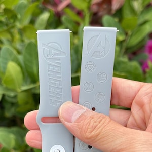 May include: Two gray silicone watch bands with different designs. One band has the word "Avengers" printed on it. The other band has various Marvel superhero logos printed on it.