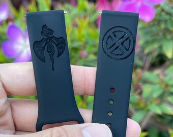 Op de afbeelding: Twee zwarte horlogebandjes met verschillende ontwerpen. Eén bandje heeft een afbeelding van een gestileerd figuur met vleugels. Het andere bandje heeft een afbeelding van een cirkel met een X erin.