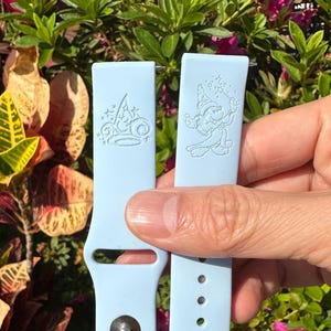 May include: Two light blue watch bands with engraved designs. One band features a crown design, and the other has a cartoon character. The bands are held up against a background of green and red foliage.