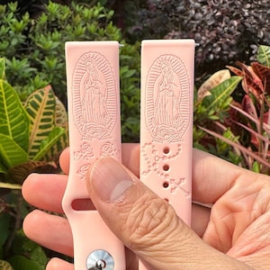 Puede incluir: Dos correas de reloj de silicona rosa con un diseño religioso. Una correa presenta una representación de la Virgen María con rosas, mientras que la otra correa presenta un rosario con una cruz.