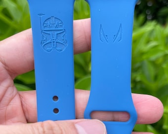 Op de afbeelding: Twee blauwe horlogebandjes met gegraveerde ontwerpen. Eén bandje heeft een helmontwerp en de andere een gestileerd embleem. De bandjes worden door een hand vastgehouden tegen een achtergrond van groen gebladerte.