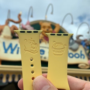 May include: Two yellow Apple Watch bands with Winnie the Pooh designs. One band features Winnie the Pooh and the other features a honey pot with the word "Hunny".