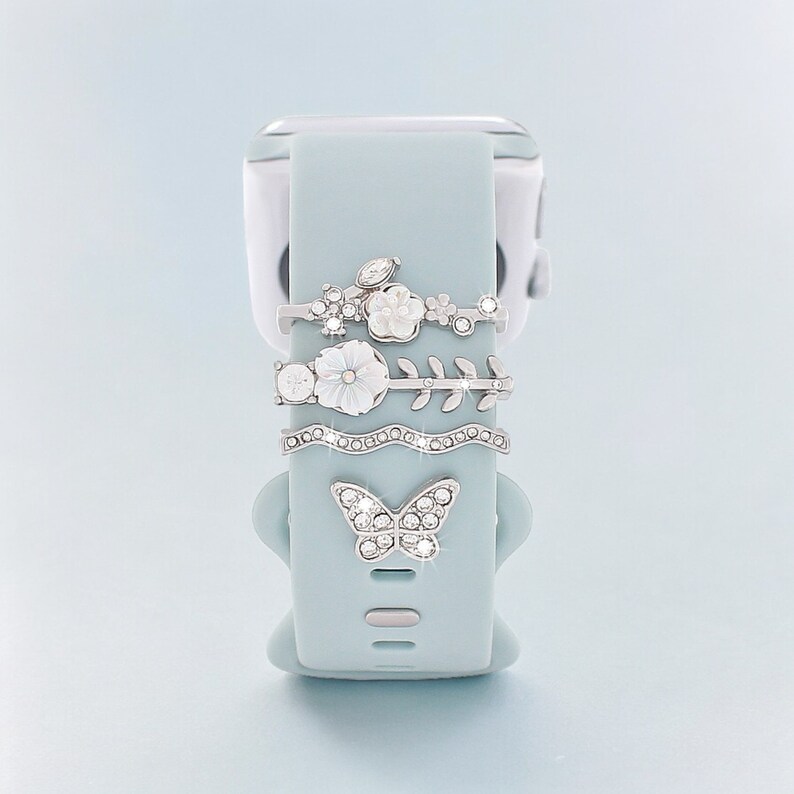 Pode incluir: Uma pulseira de rel&oacute;gio de silicone azul claro com um amuleto de borboleta prateado e tr&ecirc;s amuletos prateados com flores brancas e strass.